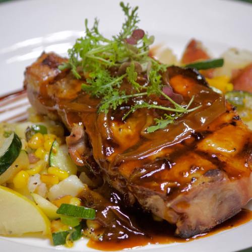 A plated dish featuring grilled meat with sauce, garnished with herbs, served with corn and vegetables on the side.