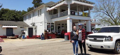 A couple stands in front of a white, two-story building with a porch and red accents, parked truck nearby.