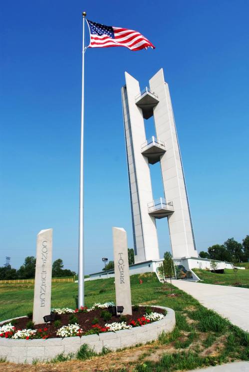 A tall, modern tower with an American flag in front, surrounded by landscaped flowers and stone markers.