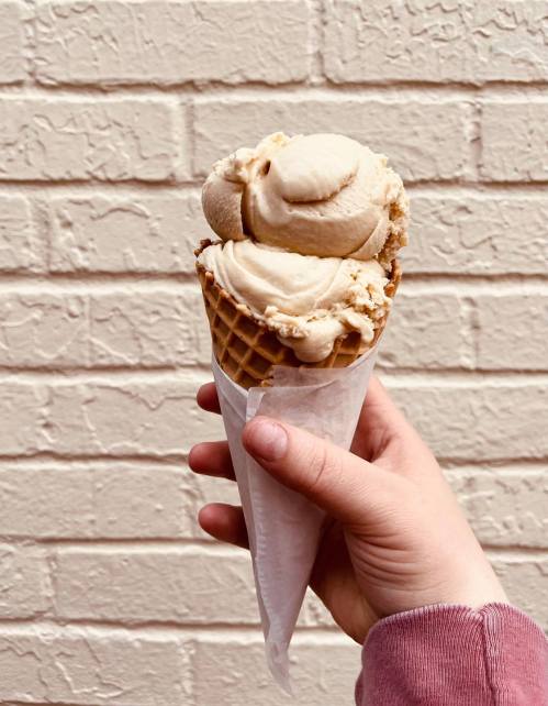 A hand holding a waffle cone with two scoops of creamy ice cream against a textured white wall.