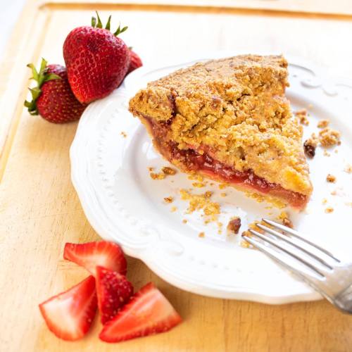 A slice of strawberry pie on a white plate, with fresh strawberries and a fork beside it on a wooden surface.