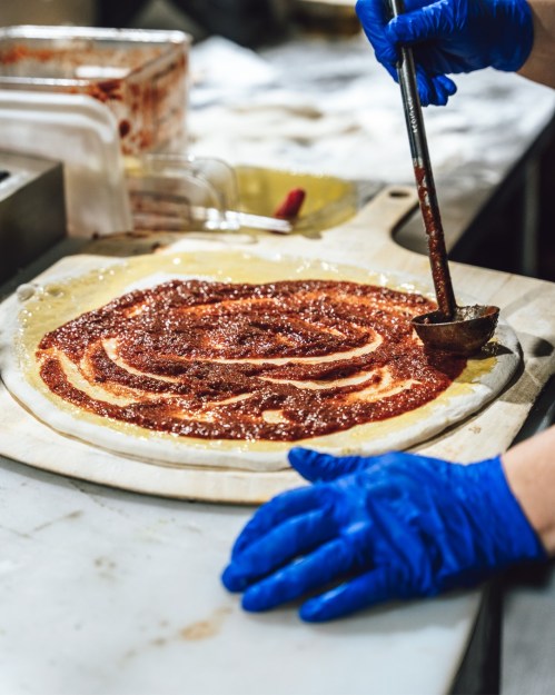 A person in blue gloves spreads tomato sauce on a pizza dough on a wooden surface in a kitchen.