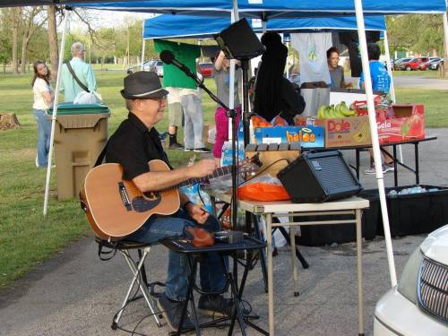 A musician with a guitar sits under a blue tent at an outdoor market, surrounded by tables of food and people in the background.