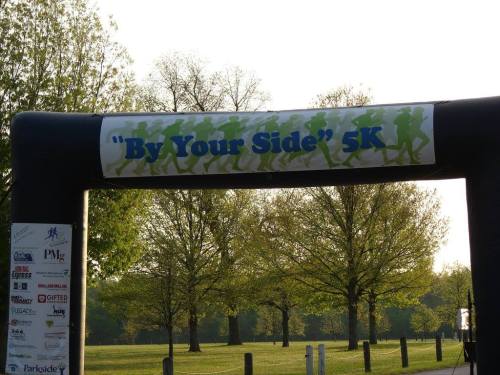 Archway for the "By Your Side" 5K event, surrounded by trees and a clear sky.