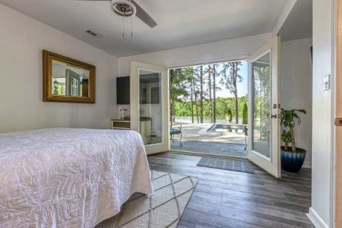 Bright bedroom with a large window opening to a patio, featuring a bed, mirror, and greenery outside.