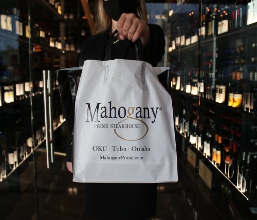 A person holding a white takeout bag from Mahogany Prime Steakhouse in front of a wine display.