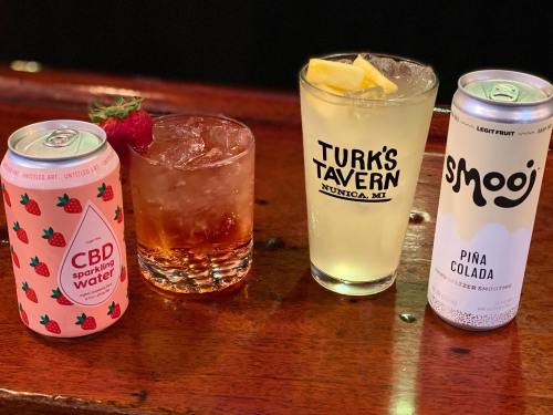 Four beverages on a wooden bar: a can of CBD sparkling water, a cocktail, and two cans of flavored drinks.