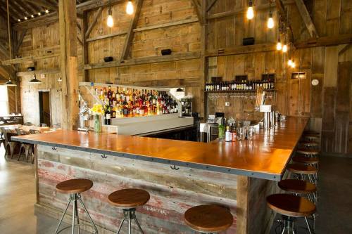 A rustic bar with a wooden counter, shelves of liquor, and pendant lights in a cozy barn setting.