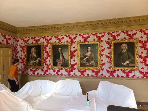 A person stands near a wall adorned with four ornate portraits, surrounded by covered furniture in a room with floral wallpaper.