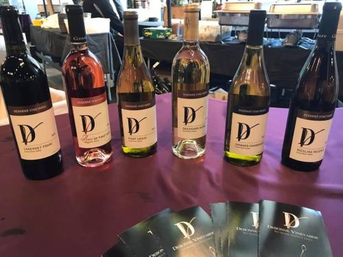 A display of six wine bottles with elegant labels on a table, alongside promotional materials.