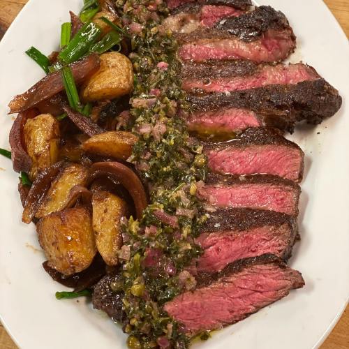 Sliced steak topped with green sauce, served with roasted potatoes and sautéed onions on a white plate.