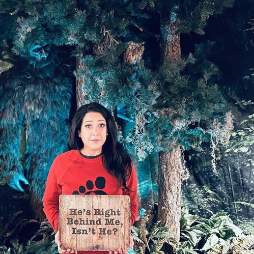 A woman in a red sweater holds a sign reading "He's Right Behind Me, Isn't He?" with a large creature lurking in the background.