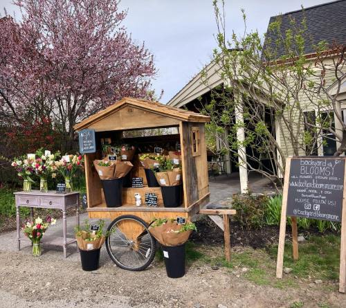 A wooden flower cart with bouquets and plants outside a charming shop, surrounded by blooming trees and greenery.