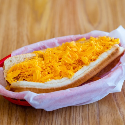 A hot dog in a bun topped with a generous amount of shredded cheese, served in a red basket with pink paper.