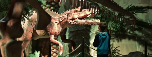 A child gazes at a large dinosaur exhibit in a museum, surrounded by lush greenery and dinosaur bones.