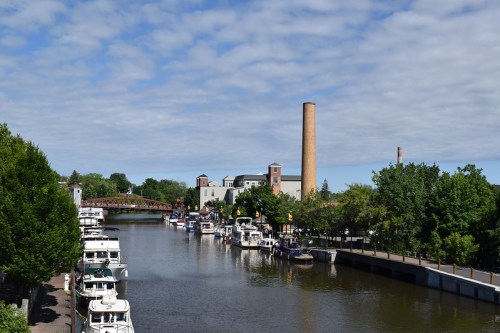 A scenic view of a river lined with boats, trees, and a tall smokestack under a partly cloudy sky.