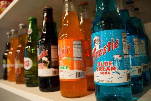 A variety of colorful soda bottles lined up on a shelf, featuring Frostie Blue Cream Soda prominently in the foreground.