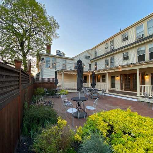 A cozy outdoor patio with tables and chairs, surrounded by greenery and a two-story building in the background.