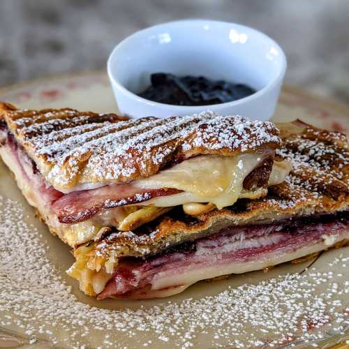 A layered pastry filled with cheese and ham, dusted with powdered sugar, served with a small bowl of jam.