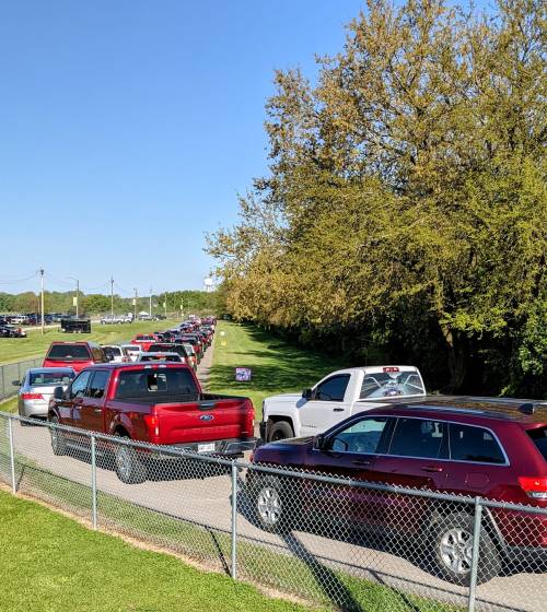 A long line of vehicles, mostly red trucks, waiting along a grassy area under a clear blue sky.