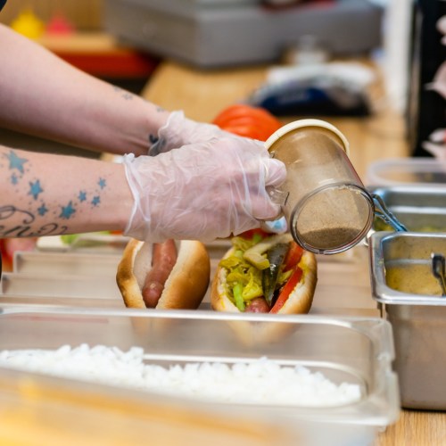A person in gloves adds seasoning to a hot dog topped with vegetables, with other ingredients visible in the background.