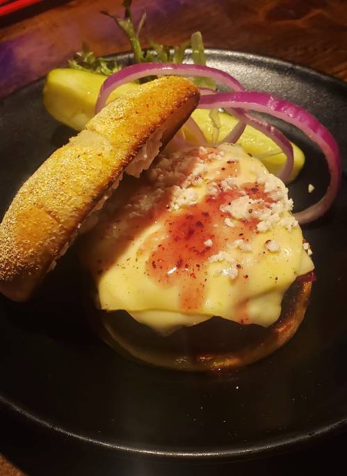 A gourmet burger with melted cheese, topped with sauce, served with pickles and red onion on a black plate.