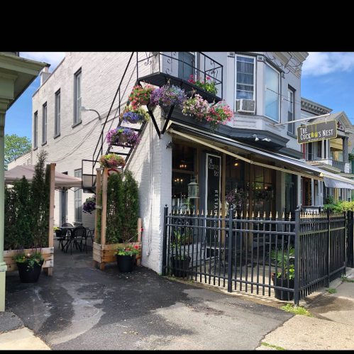 A charming restaurant exterior with flower baskets, outdoor seating, and a black fence in a quaint neighborhood.