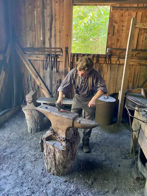 A blacksmith works at an anvil in a rustic workshop, surrounded by tools and wooden structures.