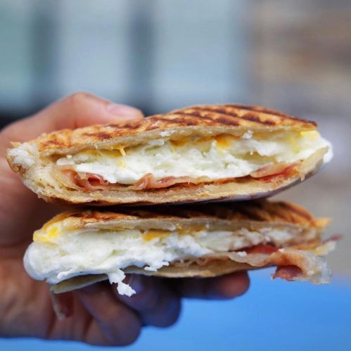 A hand holding a toasted sandwich filled with eggs, cheese, and ham, showcasing its layers.