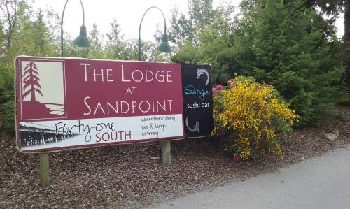Sign for "The Lodge at Sandpoint" featuring a sushi bar, surrounded by greenery and colorful flowers.