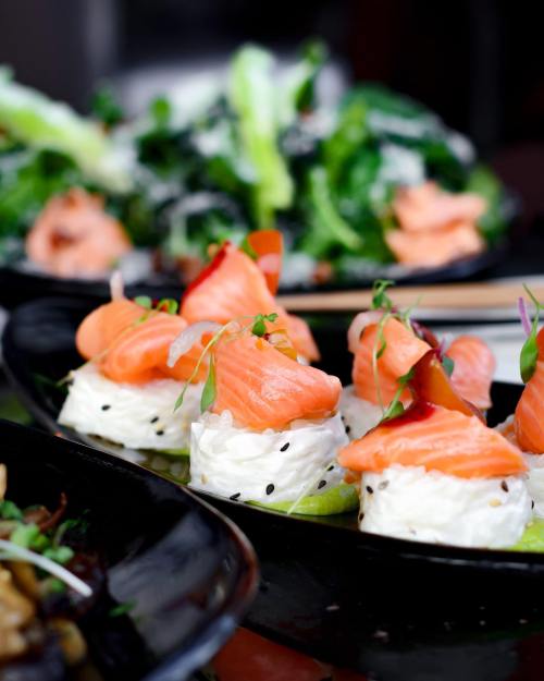 A close-up of sushi rolls topped with salmon and garnished with microgreens, served on a black plate.