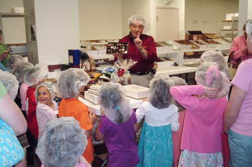 A group of children in hairnets listens attentively to an adult in a red shirt, surrounded by candy boxes and treats.