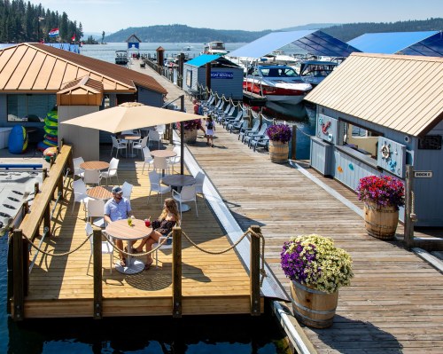 A couple enjoys drinks on a sunny dock with boats and colorful flowers in the background by a lake.