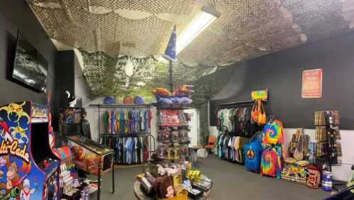 A colorful shop interior with clothing racks, bags, and an arcade game under a camo net ceiling.