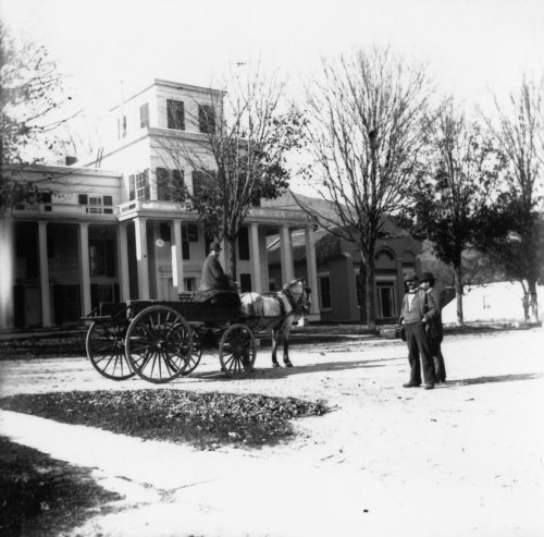 A horse-drawn carriage in front of a large building, with two men standing nearby among bare trees.