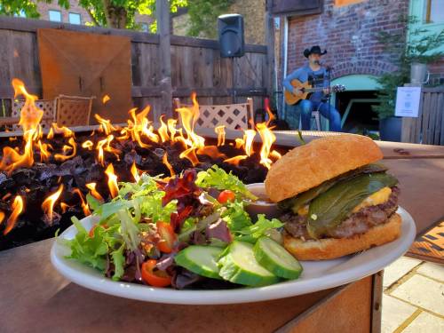 A delicious burger with greens and salad, with a fire pit and a musician playing guitar in the background.