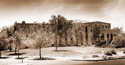 Historic building surrounded by trees, captured in sepia tones, with a person standing on the sidewalk in the foreground.
