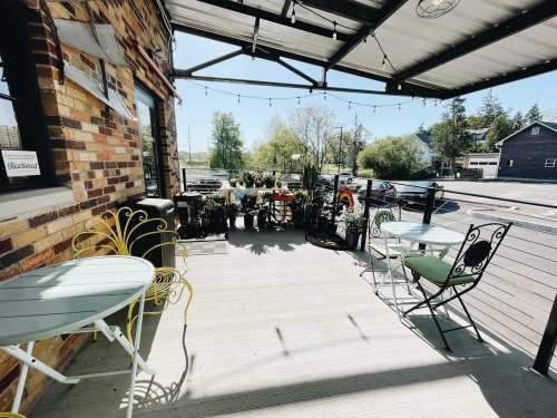 A cozy outdoor patio with two small tables and chairs, surrounded by greenery and string lights.