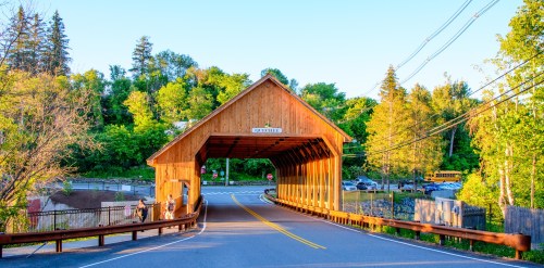 A wooden covered bridge spans a road, surrounded by lush green trees and a clear blue sky.
