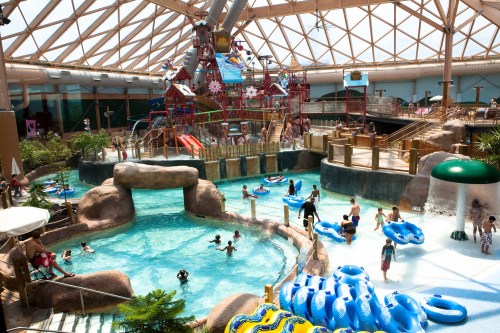 Indoor water park featuring a pool, water slides, and children playing with inflatable tubes under a glass roof.