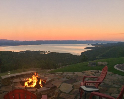 A serene sunset over a lake, with a fire pit and red chairs in the foreground, surrounded by lush greenery.