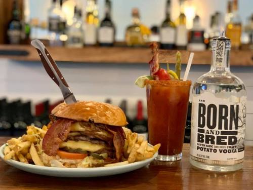 A juicy burger with bacon and fries, alongside a cocktail and a bottle of potato vodka on a bar counter.