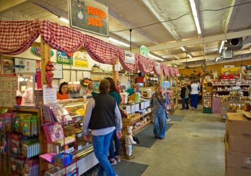 A bustling market with colorful stalls selling pies, cider, and various goods, filled with shoppers and cheerful decor.