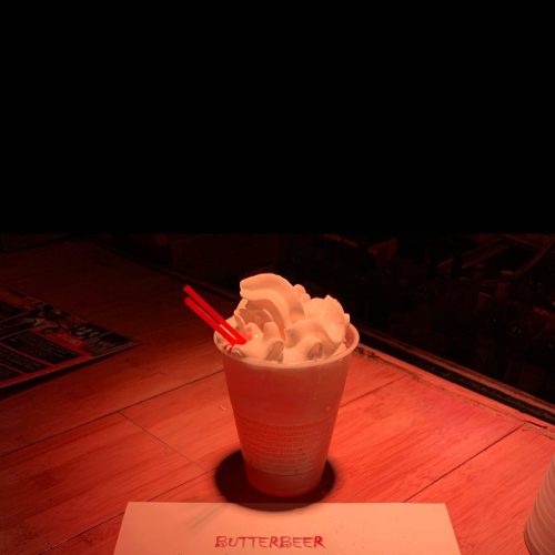 A cup of butterbeer topped with whipped cream and two red straws, sitting on a wooden surface.