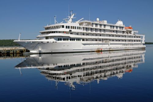 A large white cruise ship docked at a pier, reflecting in calm blue water under a clear sky.