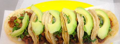 Five tacos filled with meat, onions, and cilantro, topped with slices of avocado, served on a yellow plate.