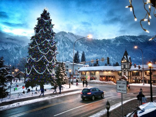 A snowy street scene with a large decorated Christmas tree, mountains in the background, and festive lights.