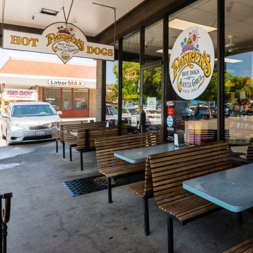 Exterior of a hot dog stand with wooden tables and chairs, featuring a sign that reads "Parker's Hot Dogs."