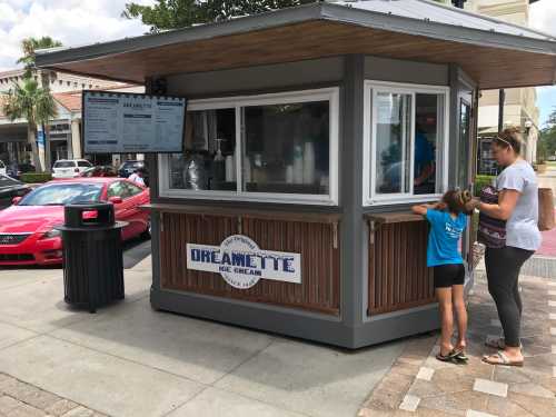 A small ice cream kiosk with a menu, where a woman and child are ordering ice cream on a sunny day.