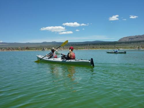 Two people kayak on a calm lake under a clear blue sky, with mountains in the background.